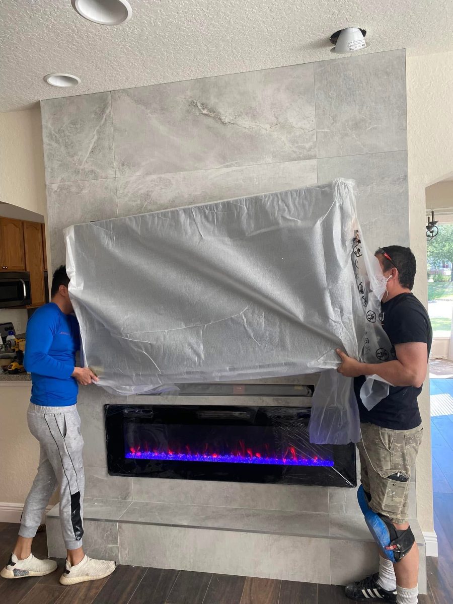 Two installers wrapping a large electric fireplace insert with protective plastic sheeting on a tiled wall