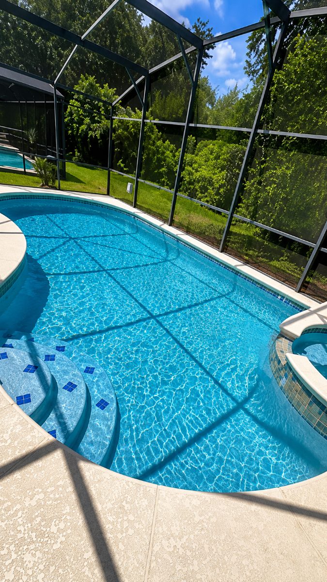 Crystal clear swimming pool with built-in spa, screened enclosure, and lush green landscaping