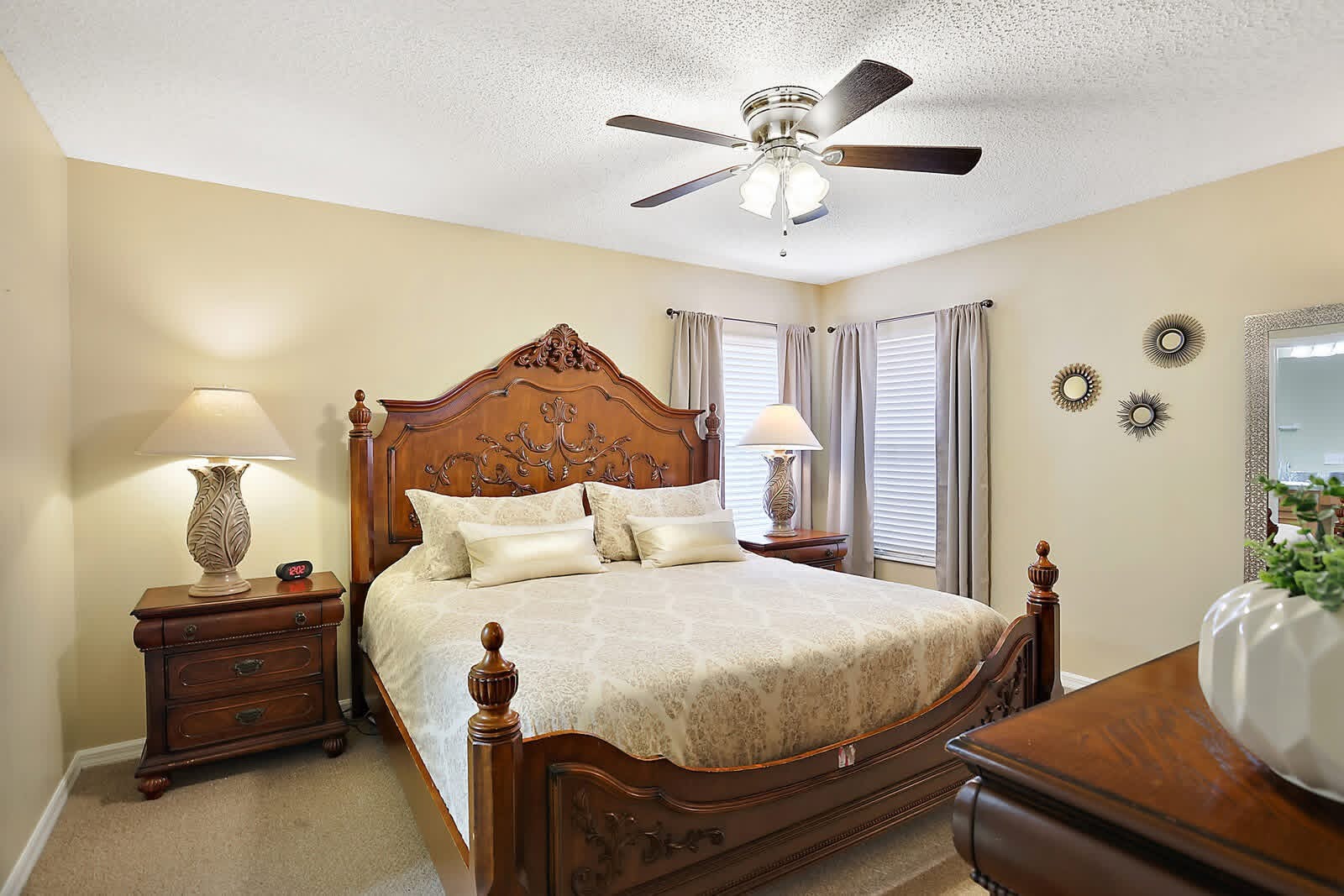 Elegant master bedroom with ornate wooden bed, cream bedding, matching nightstands, ceiling fan, and cream-colored walls